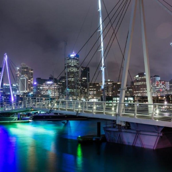 Wynyard Crossing bridge lit up at night above the water with views of yachts and boats in the harbour and the Sky Tower and sky scrapers lit up